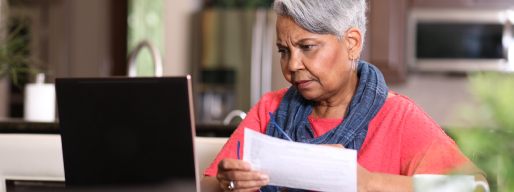 Senior adult woman at home sitting at kitchen table paying bills or online banking using laptop computer. She is visibly frustrated. Paperwork and coffee cup on table.