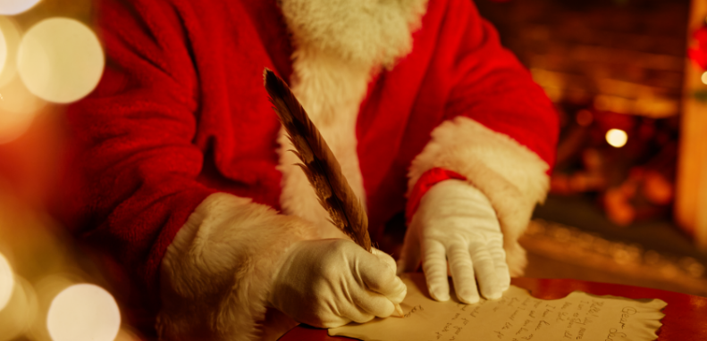 Close up of man with a white beard, wearing a red suit with white trim writing a letter. 